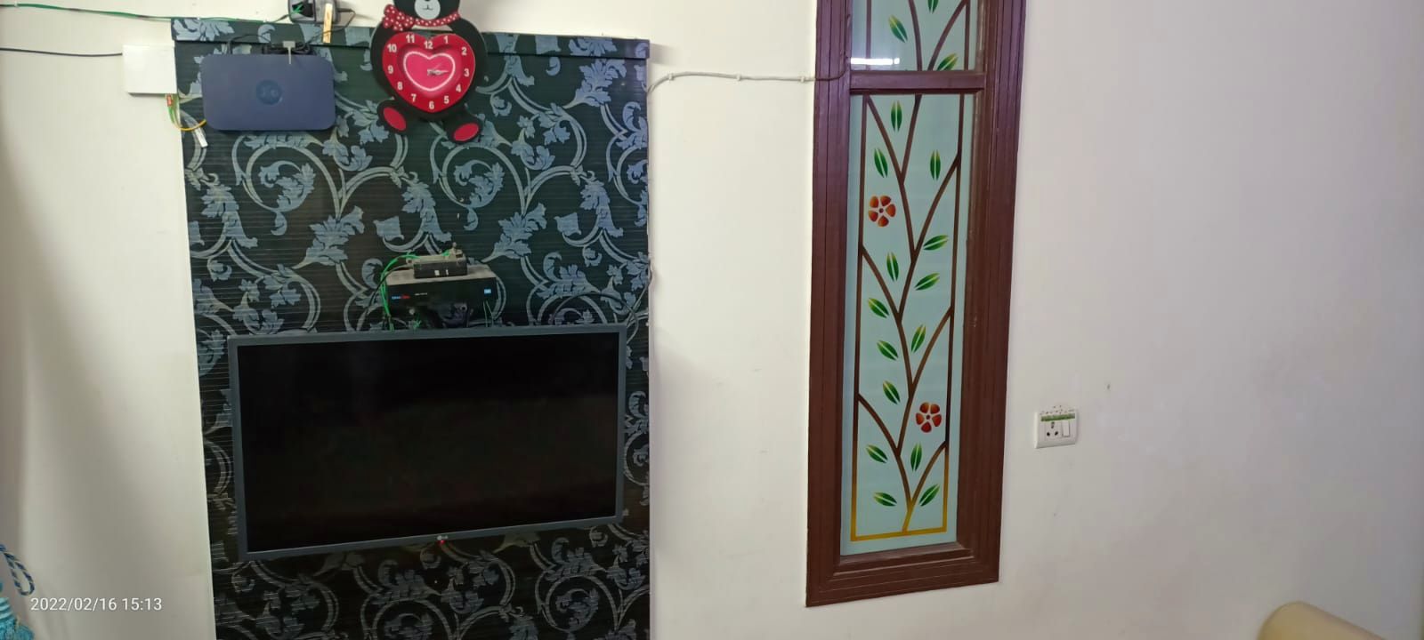 The living room before work: a television on a plain wall with dark patterned wallpaper and exposed cabling.